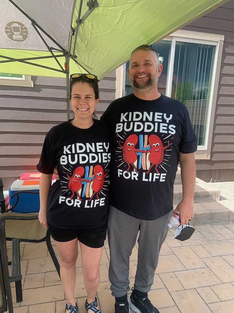 Outside on a patio, Josh Raymond is pictured with his kidney donor Shiloh. They wear matching black cartoon kidney shirts with the text Kidney Buddies for Life. Shiloh has light skin, brown eyes, and curly brown hair. Josh is a head taller than Shiloh and has light skin, blue eyes, short gray hair, and a gray beard. In the lower right corner of the photo is a logo that says Touched by Transplant with flowering green and teal vines.