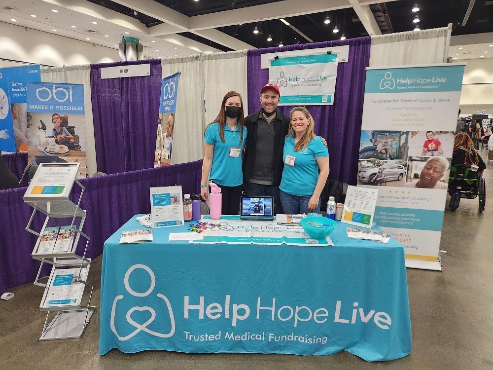 At the Help Hope Live booth at Abilities Expo LA, which is branded with teal and the tagline Trusted Medical Fundraising, Karli and Sonny are with client Ambassador Dylan Mortimer. Karli has light skin, straight brown hair, and a black face mask and she wears a teal Help Hope Live polo. A head shorter than Karli, Sonny has light skin, sandy straight hair, and a teal Help Hope Live polo. Half a head taller than Karli, Dylan has light skin, a red ball cap, and a charcoal gray sweater.