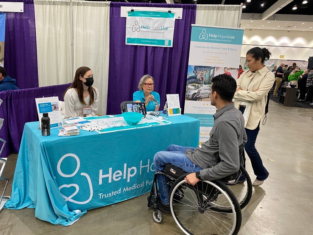 Help Hope Live's Karli and client Ambassador Rachelle Ledbetter are seated behind the teal Help Hope Live table at Abilities Expo LA 2024. The table is covered with brochures, pens, and other materials. Karli has light skin, a white top, straight brown hair, and a black face mask. Rachelle has light skin, glasses, shoulder-length gray hair, and a teal Help Hope Live polo. In front of the table, a man in a wheelchair is speaking with Rachelle. He has brown skin and short black hair. A woman listening beside him has brown skin, curly black hair, and a cream collared shirt.