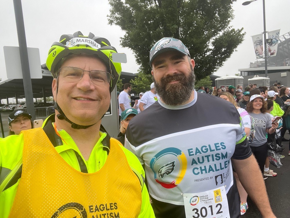 Heart transplant recipient Bill Soloway takes a selfie with Eagles football player Jason Kelce at an outdoor event for Eagles Autism Challenge. Bill has light skin, glasses, and bright yellow-green cycling gear and a helmet with an Eagles Autism Challenge gold bib overtop. Jason Kelce has light skin, his signature full brown beard, and an Eagles ball cap with a shirt that reads Eagles Autism Challenge.