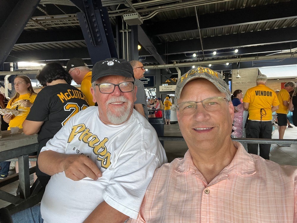 Heart transplant recipient Bill Soloway takes a selfie with his heart donor's father in the stands of a Pittsburgh sports event. Bill has light skin, glasses, and a ball cap. His donor's father has light skin, a white mustache and goatee, glasses, and a ballcap.