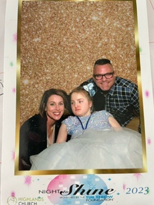 In souvenir photo from the Highlands Church Night to Shine event in 2023 sponsored by the Tim Tebow Foundation, Reese D'Ortenzio is seated in a mobility device and wears a sparkling light blue and tuille dress. She has a matching light blue bow in her hair and she has light skin and fair hair. Her mom beside her has light skin and short dark hair. Her dad has light skin, black glasses, and short dark hair.