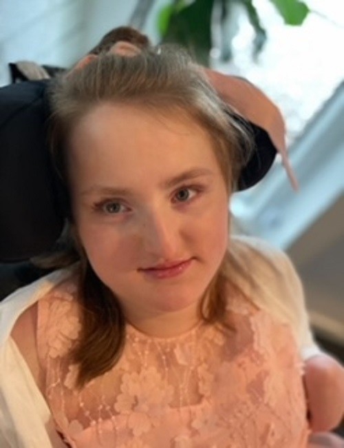 In a portrait, teenager Reese D'Ortenzio wears a light pink beaded and floral formal dress and sits in her black mobility device. Reese has light skin, shoulder-length brown hair, and blue-gray eyes.