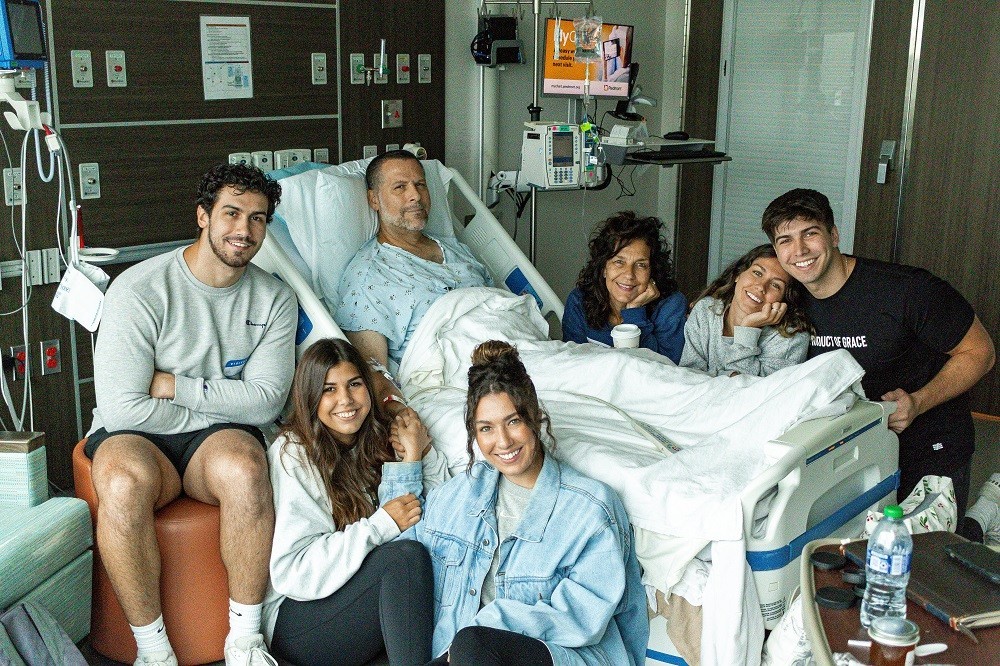 Heart transplant recipient John A Lee is in a hospital bed surrounded by his wife and five adult children. John has light skin, short dark hair, and a short gray beard.