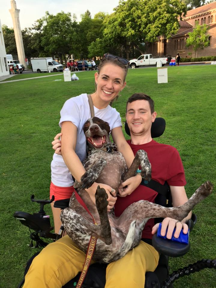 Help Hope Live client Dillon Connolly with his large service dog on his lap as he sits in his power chair outside.