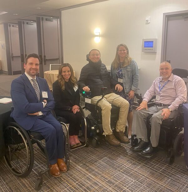 Sonny stands beside two individuals seated in manual wheelchairs and two individuals seated in power chairs. Two of the individuals are Kelley Simoneaux, who has a black suit, striking pink socks, and sandy-colored wavy hair with light skin and red lipstick, and Josh Basile, client Ambassador for Help Hope Live, with light skin, short brown hair, brown eyes, a black puffer jacket and scarf, and khaki pants.
