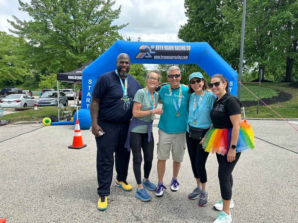 Five current and former Help Hope Live supporters gather in front of the Bryn Mawr Racing Co finish line at Hope Travels 2024. From left to right: Barrett Brooks, who has brown skin and a short black and gray goatee, former Executive Director Lynne Samson, who has light skin with gray hair and glasses, board member Ron Siggs, who has light skin, short white hair, and a Hope Travels teal t-shirt, founding volunteer Chris Kanter, who has light skin and shoulder-length dark hair, and current Executive Director Kelly L Green, who has light skin, dark hair, sunglasses, and a rainbow tutu.