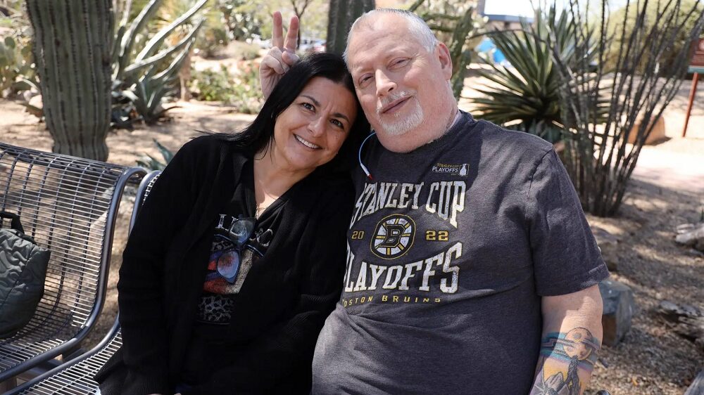 Larynx transplant recipient Marty Kedian gives his wife Gina bunny ears as they both sit on a park bench in a desert environment.