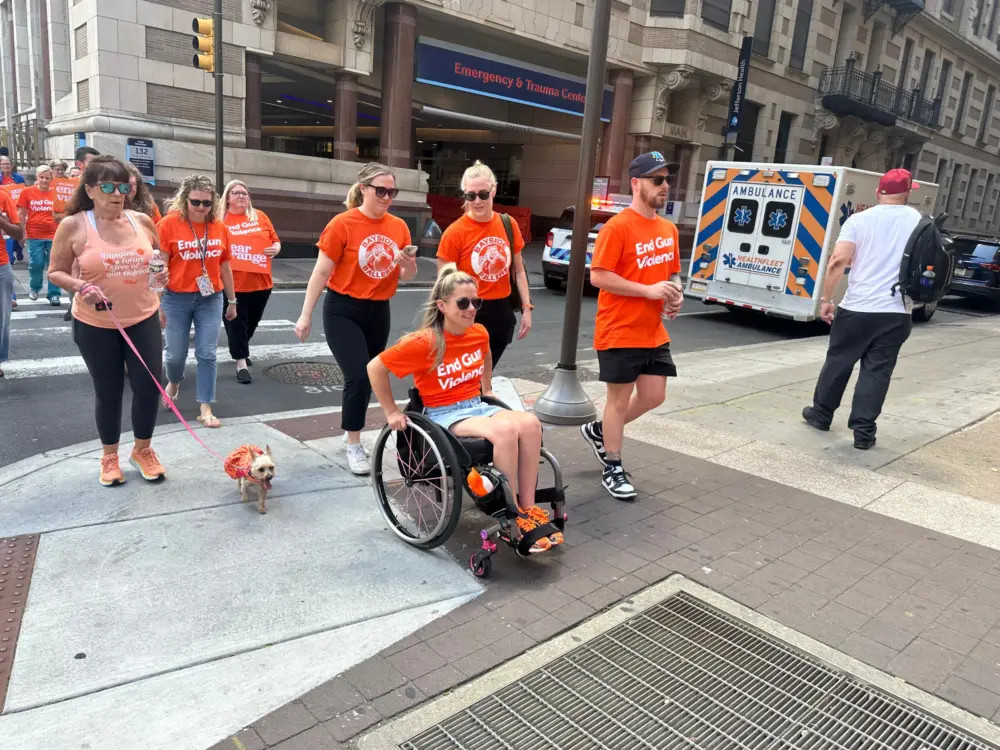 38-year-old Amanda Parezo is seated in her manual wheelchair on a Philadelphia city street wearing orange sneakers and an orange shirt that reads End Gun Violence. She has light skin, blonde hair, and sunglasses. A group of 10 people walking behind her all wear the same t-shirt.