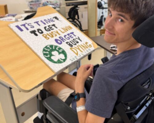 Drew Boniface seated in his wheelchair in rehabilitation with a sign that says It's Time to Get Busy Livin or Get Busy Dying with a Celtics hand-drawn logo.