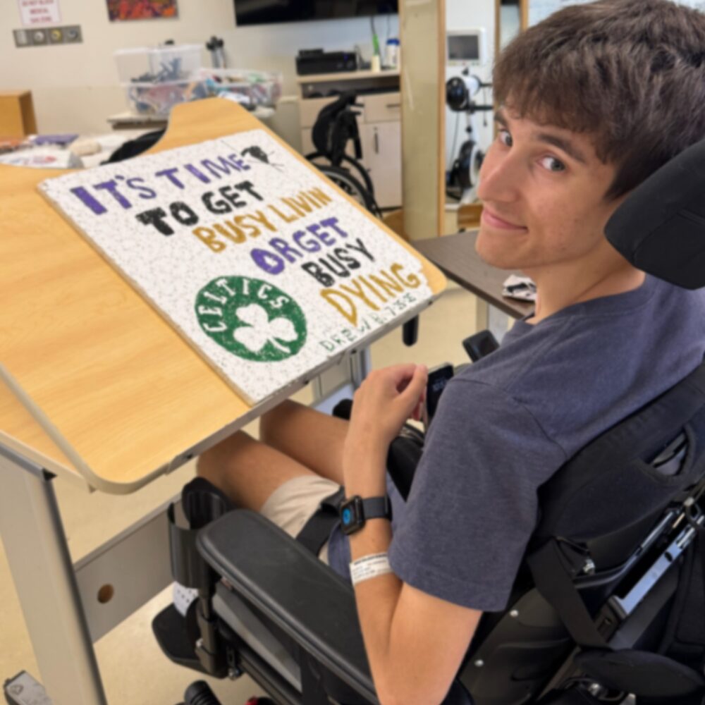 Drew Boniface seated in his wheelchair in rehabilitation with a sign that says It's Time to Get Busy Livin or Get Busy Dying with a Celtics hand-drawn logo.