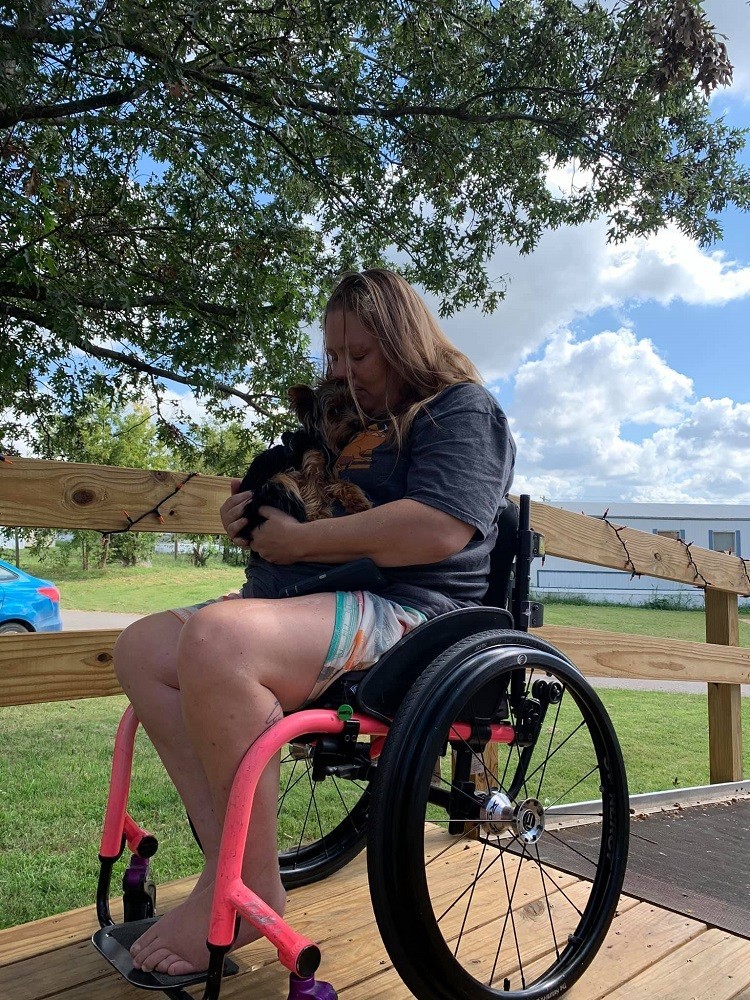 Help Hope Live client Jamie Carlson aka the "Cerebral Palsy Quilter" is seated in her wheelchair on a deck cradling a terrier.
