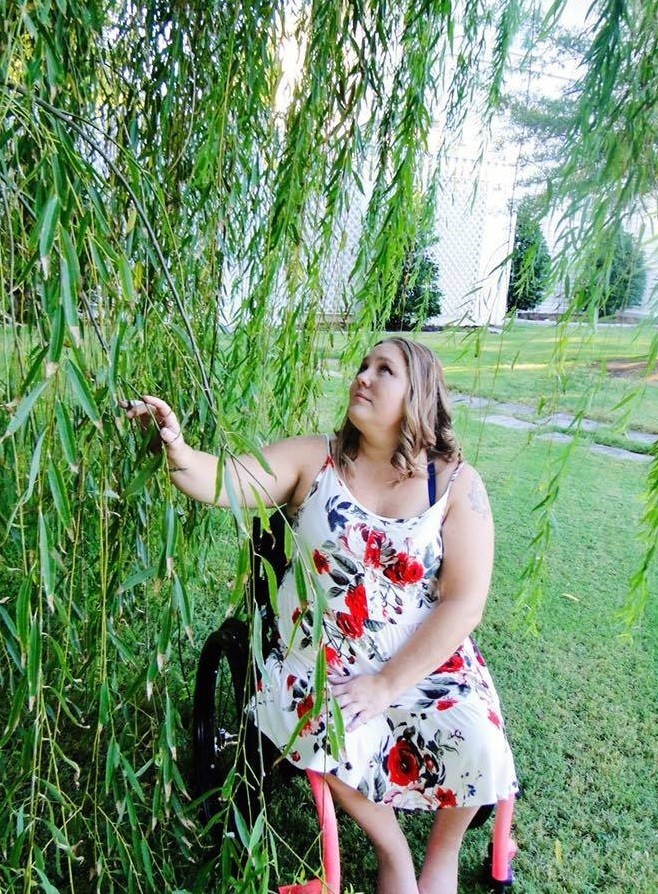 Help Hope Live client Jamie Carlson aka the "Cerebral Palsy Quilter" is outside in her manual wheelchair reaching for the branches of a weeping willow.