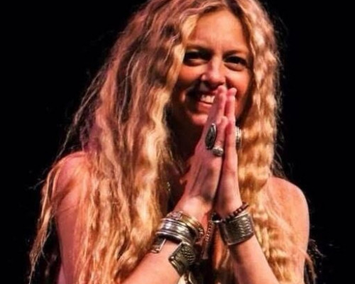 Woman with long blonde hair smiling and waving, wearing a colorful dress.