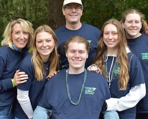 Six smiling people wearing matching navy shirts pose together outdoors among trees