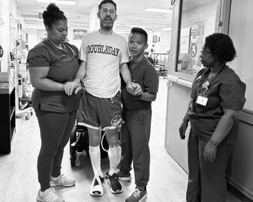 A patient wearing leg braces walks with assistance from two healthcare workers while another observes