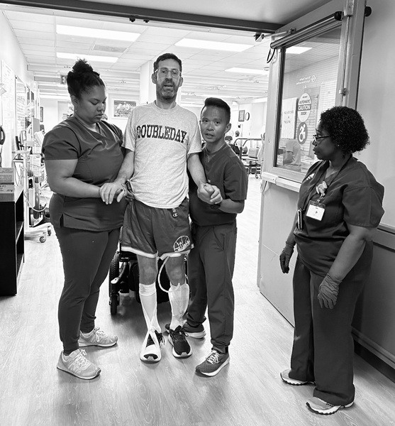 A patient wearing leg braces walks with assistance from two healthcare workers while another observes