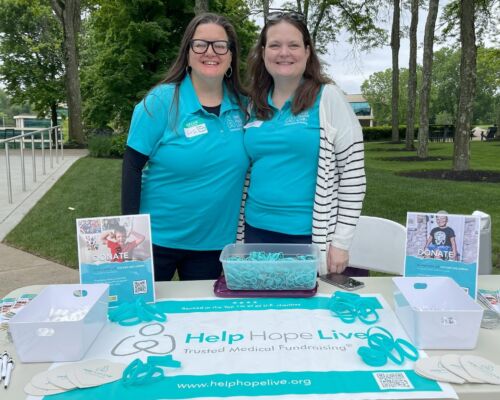 Two women staff a Help Hope Live fundraising table with donation signs, teal wristbands, and promotional materials