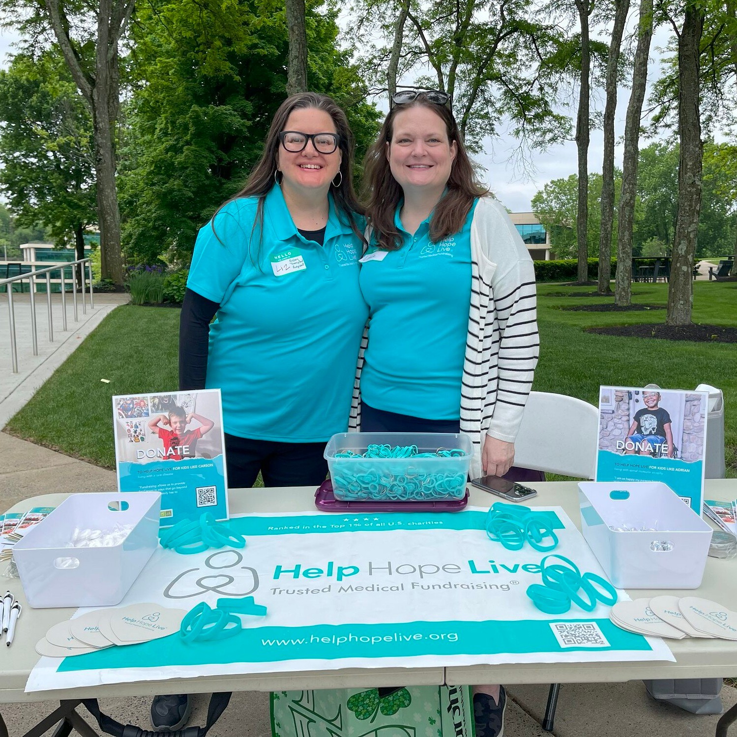 Two women staff a Help Hope Live fundraising table with donation signs, teal wristbands, and promotional materials