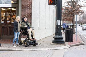 Two people at a street corner - one stands and one uses a seated mobility device that raises him to eye level.