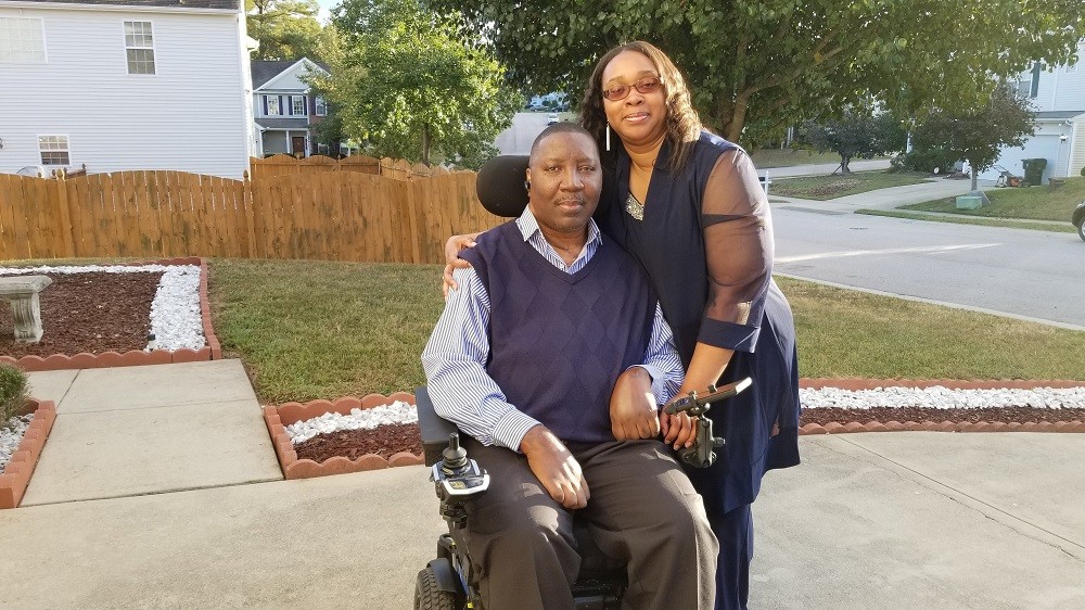 Stephany Golden is in an electric wheelchair with his wife Katrina standing beside him. They are outside.