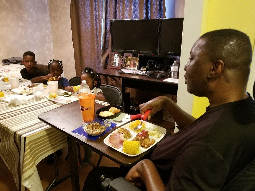 Stephany Golden is in a power chair using adaptive eating utensils. Three young children are at the dinner table across from him.