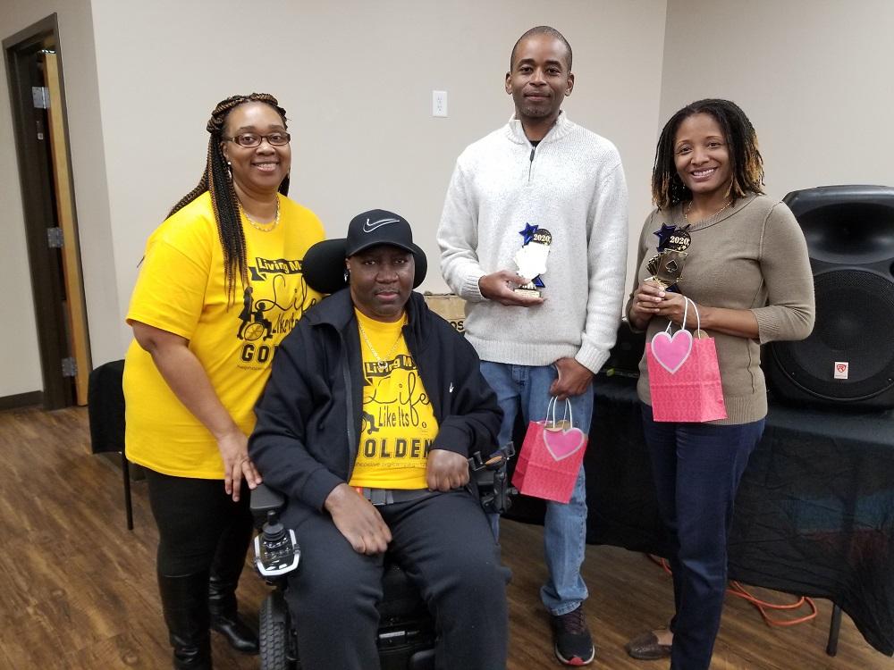 Stephany Golden and his wife Katrina are wearing matching fundraiser t-shirts in bright yellow. Stephany is in his power chair. Two supporters are standing nearby holding pink bags.