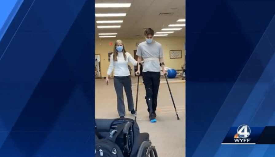 In a video screenshot from WYFF news, Austin Somerville walks with assistance with two crutches in physical therapy.