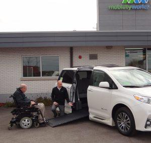 A MobilityWorks employee kneels next to a black wheelchair ramp leading into a white accessible vehicle. Beside him is an individual using a black power chair who is approaching the vehicle's ramp. They are outside a MobilityWorks building.