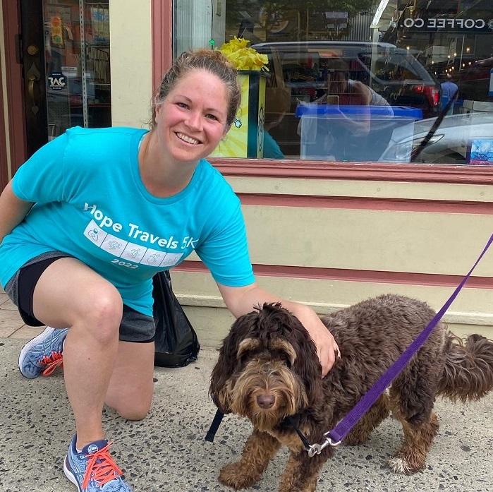 A woman kneels on one knee and smiles next to a brown curly haired dog on a purple leash.