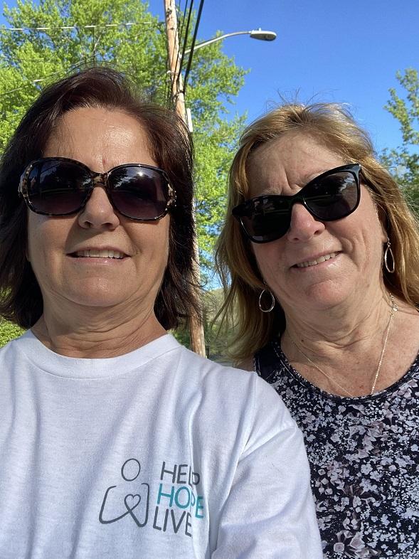 Two women wearing sunglasses smile at the camera outdoors. One is wearing a white t-shirt with the Help Hope Live logo.