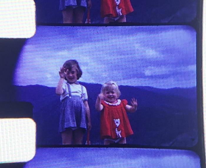 An old photograph of two children. One has short brown hair and the other has short white blonde hair. One is wearing a blue jumper and the other is wearing a red dress with a white collar. There are some low mountains and clouds visible behind them and they are waving happily at the camera.