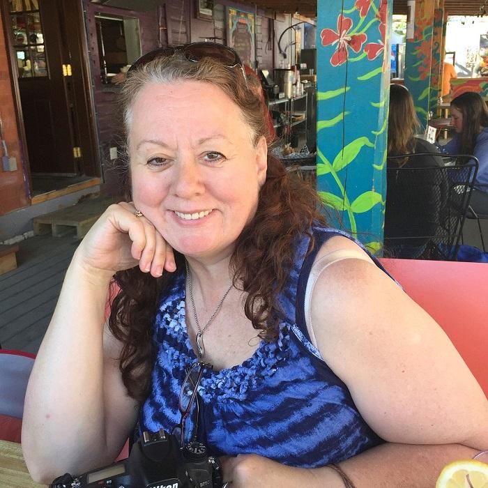 Tracey Porreca is seated in a light pink booth. She has brown eyes, curly brown hair, and a sleeveless light and dark blue striped shirt. She is smiling.