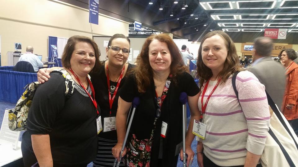 Tracey Porreca with three friends at a conference or expo. She has curly brown hair and is standing and using crutches.