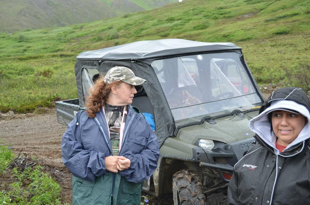 Tracey Porreca stands outside a small all-terrain vehicle with rolling green landscape visible behind her. She is wearing a camoflage ball cap and a blue windbreaker and she is looking to the right side of the photo across the top of the vehicle. She has curly brown hair. There is one other person in the frame.