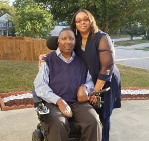 Stephany is seated in his power chair with Katrina standing beside him. They are outside of their home.