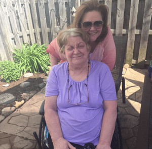 Mary Zagar is seated in her wheelchair with a purple shirt on. She has gray hair. Her daughter Patty is behind her with brown hair, sunglasses, and a pink shirt. They are outside on a patio with a fence.