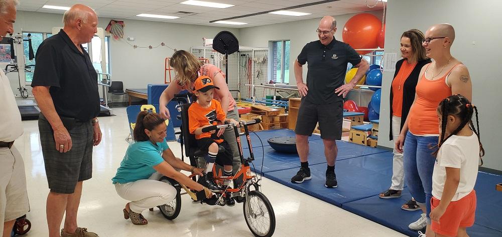 7-year-old David Albino is sitting on his new adaptive bike, which is orange and black and Philadelphia Flyers themed. He is wearing a Flyers jersey and a matching orange and black Flyers ball cap. Help Hope Live Executive Director Kelly L Green is in a teal Help Hope Live t-shirt and is bending down to help place David’s right foot on the bike pedals. Standing nearby are Bob Kelly and Brad Marsh of the Flyers Alumni Association dressed in black and David’s family dressed in orange. They are at Schreiber Pediatric Center where David receives therapy. Blue exercise mats are visible.