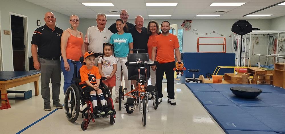 7-year-old David is in his wheelchair in a Philadelphia Flyers orange and black jersey and ball cap. His family, the Flyers Alumni Association, and Help Hope Live’s Executive Director are standing behind him and smiling. To his left is his new Flyers themed orange and black adaptive bike from Freedom Concepts. They are at Schreiber Pediatric Center where David receives therapy. Blue exercise mats are visible.