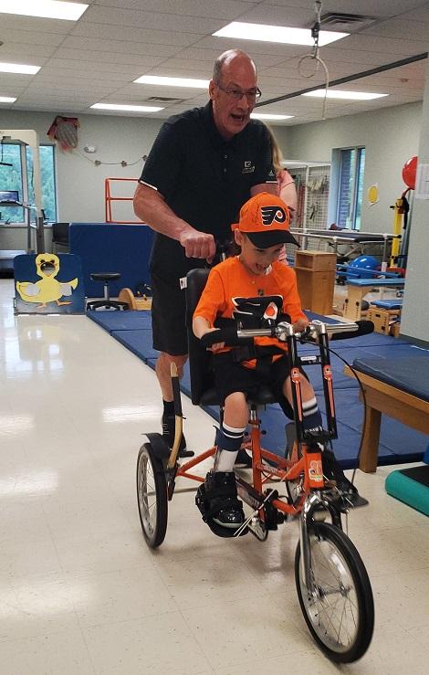 Brad Marsh of the Flyers Alumni Association excitedly pushes David on his new adaptive bike. David is wearing a Philadelphia Flyers themed bright orange and black jersey and a matching ball cap. He looks very excited and is smiling with his mouth open while looking down at his new bike. They are at Schreiber Pediatric Center where David receives therapy.