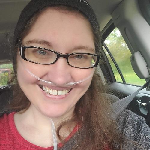 Female presenting double lung transplant recipient Bobbie has white skin with brown hair and light blue eyes. She is in a car wearing a red shirt and a gray sweater. She is wearing black square glasses and has an oxygen tube. She is smiling wide.