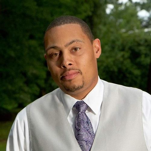 Male presenting kidney transplant recipient Lawrence has light brown skin, close cropped brown facial hair, very short styled dark hair, and dark eyes. He is wearing formal attire with a silver vest and tie.