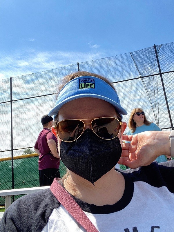 Linda Jara wears a powder blue visor with a Donate Life logo, a black mask, and brown aviator sunglasses. She is outdoors at a sporting event with a blue sky in the background.