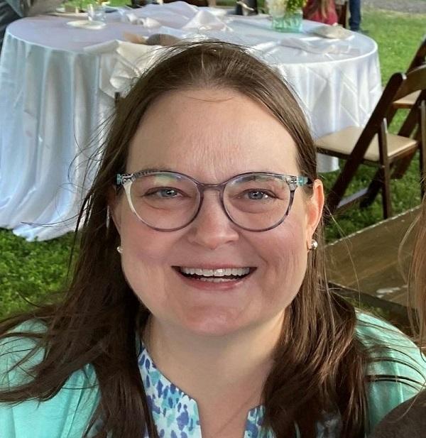 Heart transplant recipient Linda Jara smiles while attending an outdoor wedding. She has shoulder-length light brown hair, green eyes, pearl earrings, and glasses with transluscent gray frames. She is wearing pastel blues and greens.