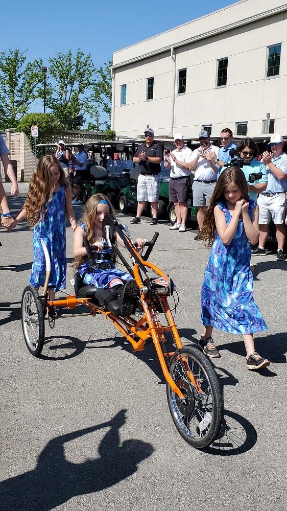 Madalynn rides her new orange Flyers-themed handcycle with her two sisters walking beside her. All three have long brown hair and are wearing blue and light blue tie-dye dresses. It’s a clear and sunny day. Behind them, a row of individuals in golf attire applaud in front of a row of golf carts.