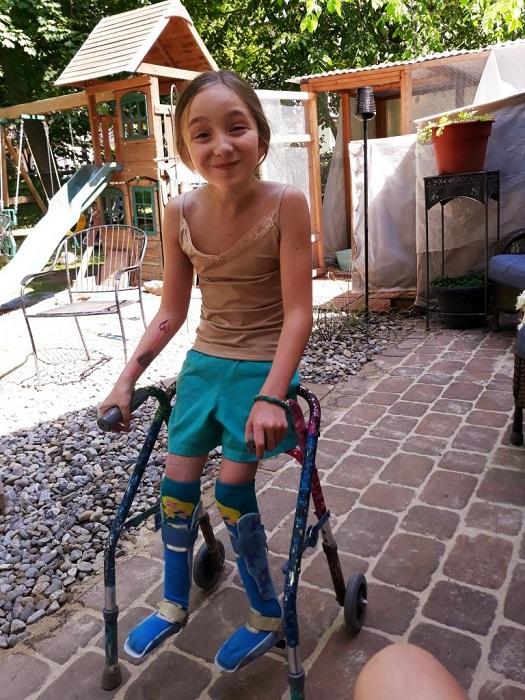 10-year-old Maddie stands in her gravel and stone paved backyard using a mobility device. Her legs are in blue over-the-knee socks and braces. She is smiling at the camera with her brown hair pulled back. She has a temporary tattoo on her right forearm, a brown tank top, and teal shorts.