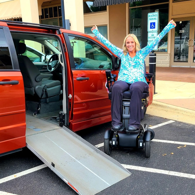 Ali Ingersoll, a woman living with paralysis, is wearing a bright aqua and white printed long-sleeved shirt and blue pants as she is seated in her black power chair next to a red accessible van with a silver ramp exended onto the blacktop of the parking lot space. She has white skin and blonde shoulder-length hair.