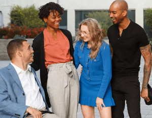 Content creators Cole Sydnor and Charisma Jamison with C Talent founder Keely Cat-Wells and an individual with brown skin in black clothing who is not named in the image. Cole has white skin and is wearing a powder blue blazer and white shirt and is seated in his power chair. Charisma is wearing a red-orange shirt and tan pants and has brown skin and short curly black hair. Keely has white skin and curly blonde hair with glasses and a blue jacket and skirt.