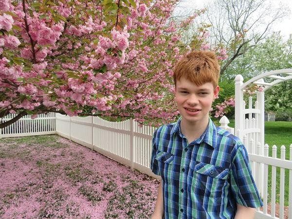 Patrick is a young teen in his Campaign Page photo. He has short red hair, light skin, and a blue and gray plaid short-sleeved button up shirt. Behind him is a tree overloaded with bright pink blossoms that have created a carpet on the yard below.