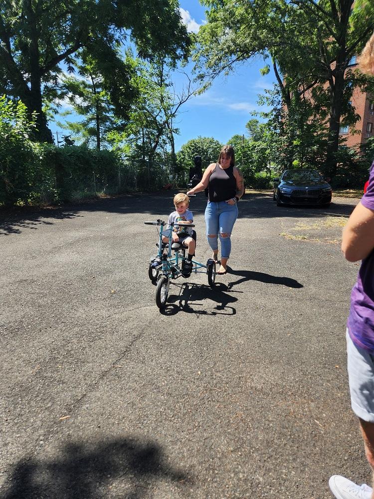 Vinny gets a push from his mom as he moves his new Freedom Concepts adaptive bike across the blacktop n front of his home. Blue sky and trees are visible in the background. Mom has light skin, brown hair with bangs, a black shirt, and jeans. Vinny is wearing shorts and a t-shirt. His new bike is teal with three black wheels.